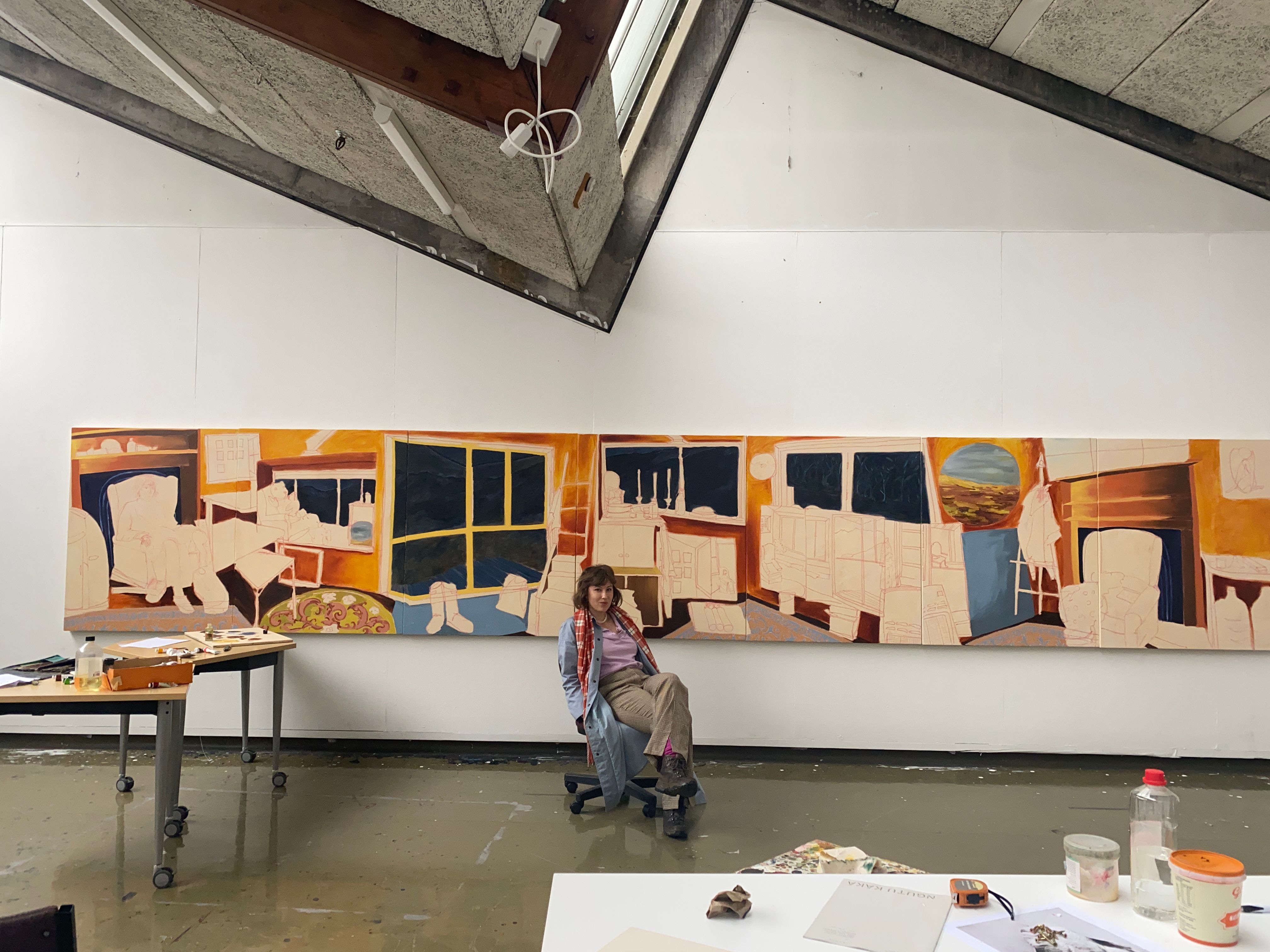 Stephanie Postles in her studio, seated in front of a long multi-panel painting.